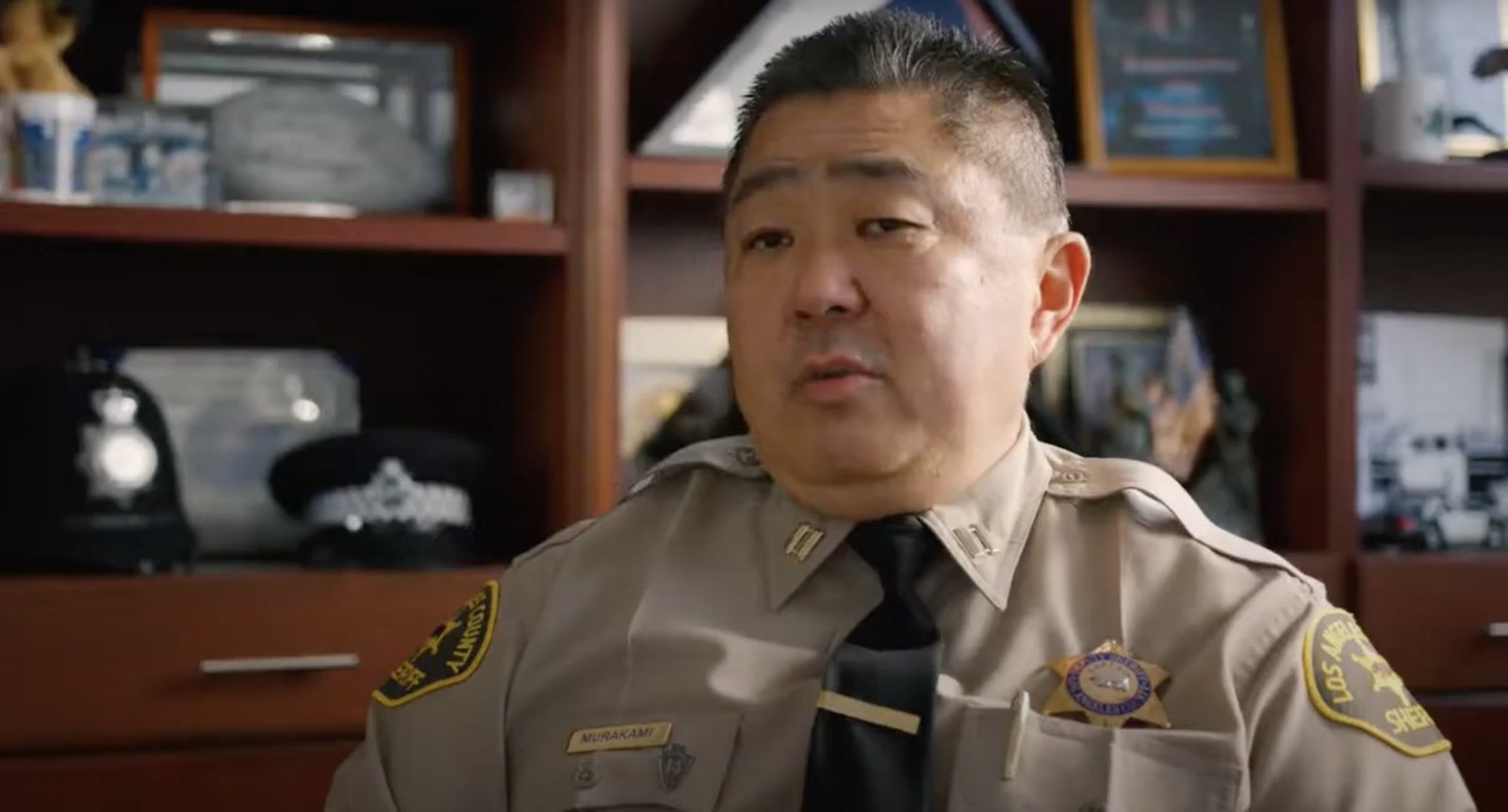 Screenshot of Timothy Murakami wearing his uniform with a shelf with police paraphernalia in the background.