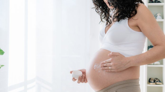 Young pretty pregnant woman with curly hair putting moisturizer on her tummy to prevent stretch marks in pregnancy.White background. copy space