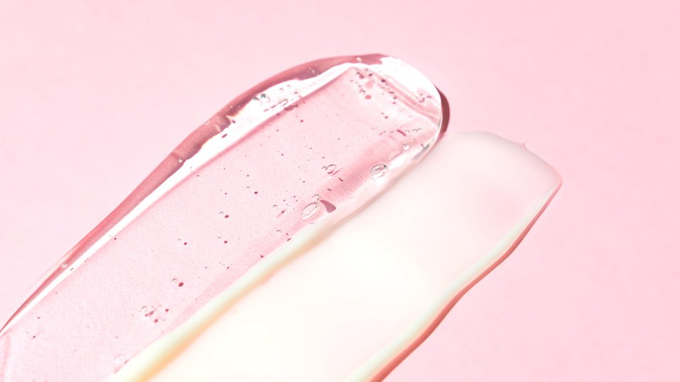 Up close flat lay shot of two serums and lotions spread out in a line on a pink background